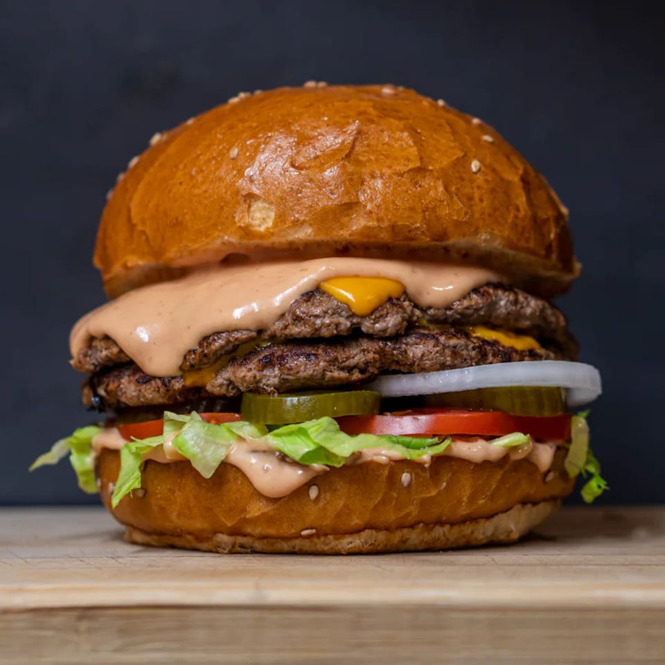 A double-patty burger with cheese, sauce, lettuce, tomato, onion, and pickles on a sesame bun.
